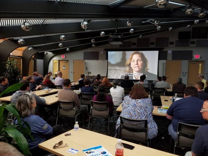 Groups of people seated at tables looking at screen with image of a woman talking.