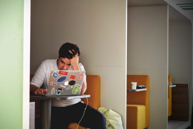 Young man with an open computer, head in hand, in a diner booth.
