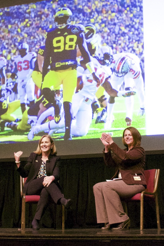 Kelly Trosvig and Cathy Curley seated on stage, image of U-M football game on projection screen in BG.