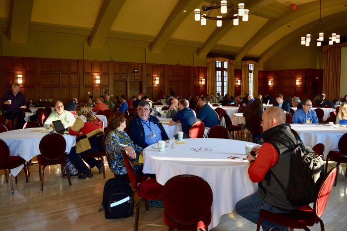 Large paneled meeting room with round tables, people seated at tables, talking.