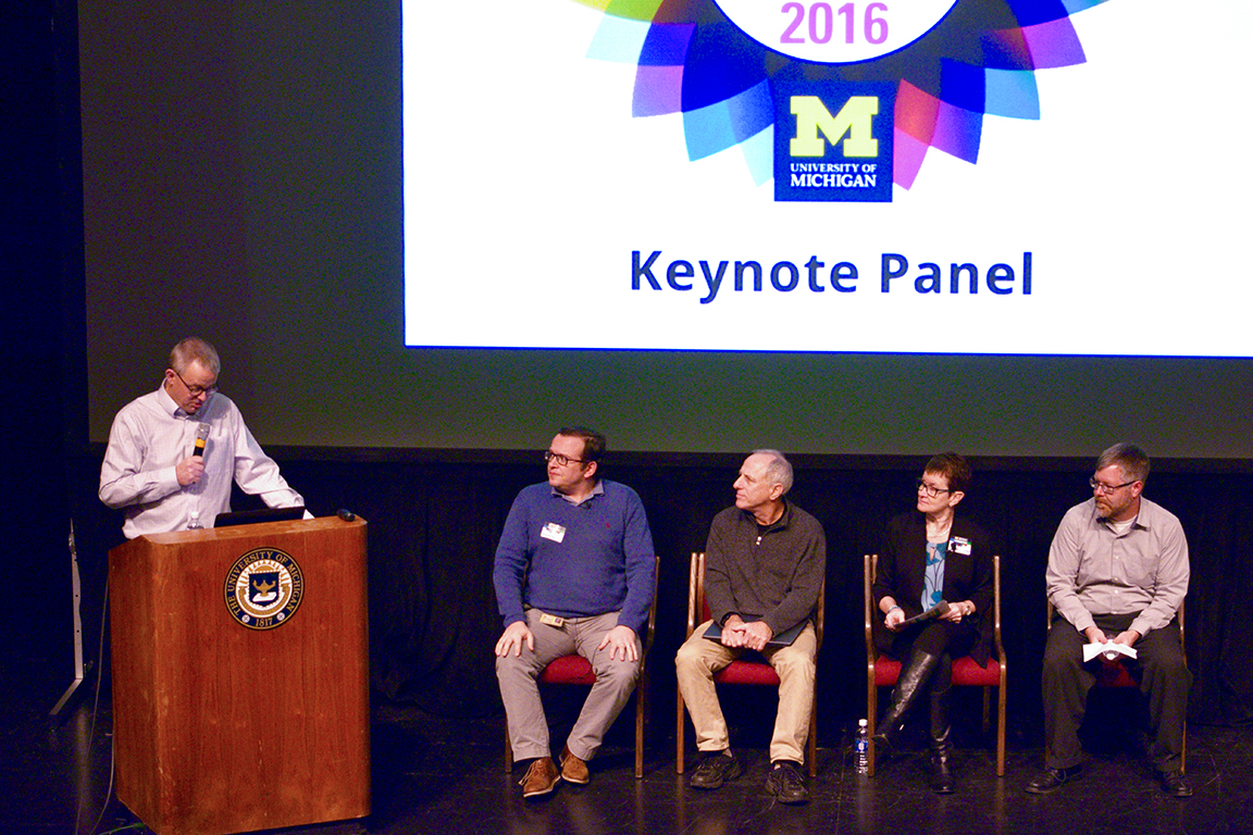 Five people on stage. One man holding mic at podium, three men and one woman seated. Slide that says Keynoe Panel with UM-logo and Symposium logo on projection screen in BG.