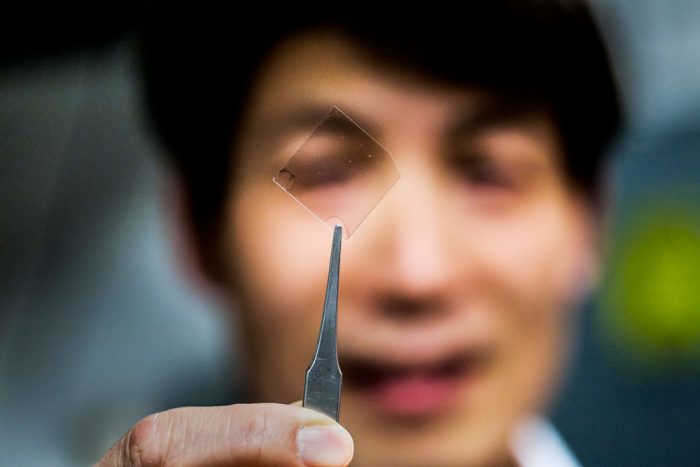 Close up photo of a piece of transparent film being held by a tweezer, soft-focus of Prof. Guo in the BG.