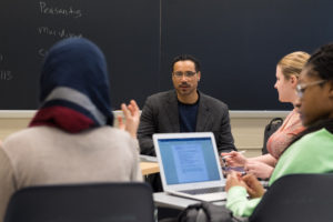 Male instructor seated in front of a blackboard with three female students.