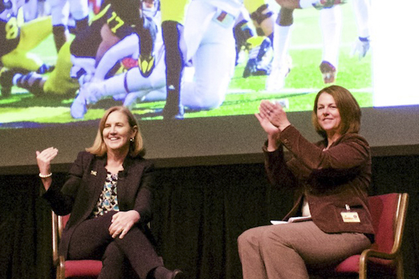 Kelly Trosvig and Cathy Curley seated on stage, image of U-M football game on projection screen in BG.