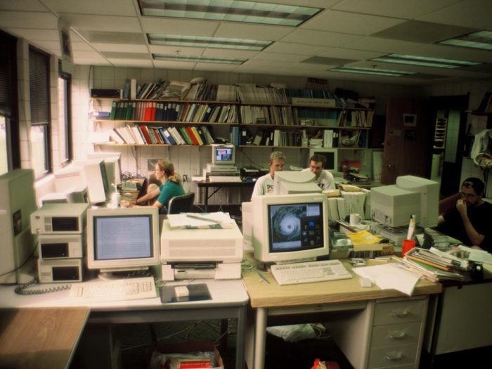 Office with computers and bookshelves, people sitting at desks, circa early 1990s.