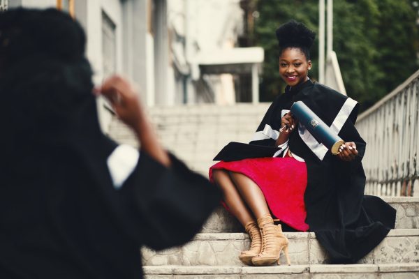 young african american woman wearing graduation robe, being photographed by another grad