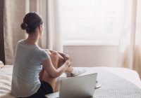 young white/asian woman sitting on bed with open laptop