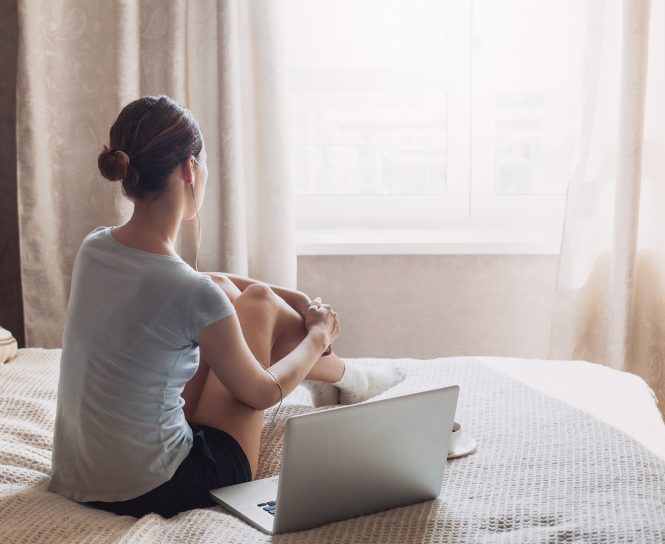 young white/asian woman sitting on bed with open laptop