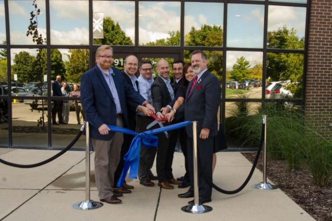 Seven people stand in front of a glass building. They standing close together and smiling while holding a giant pair of scissors moments before cutting a blue ribbon.