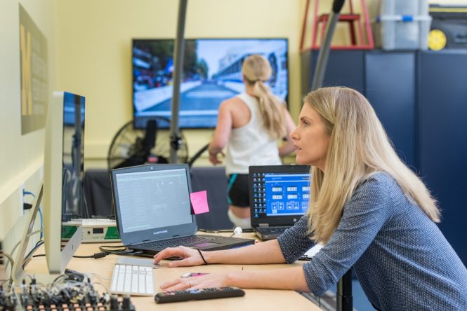 Blond woman working at computer with another woman in the background viewing a large video screen.