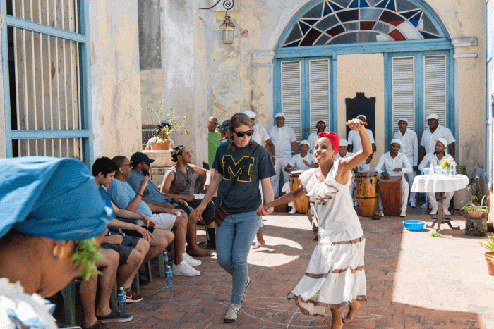Woman wearing Block M t-shirt with costumed dancer in Cuba, surrounded by tourists and musicians.