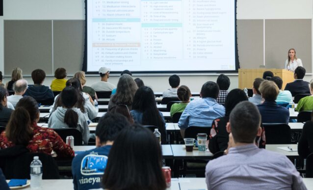 Students sitting at desks with their backs to the camera. Lecturer at the front with a presentation.