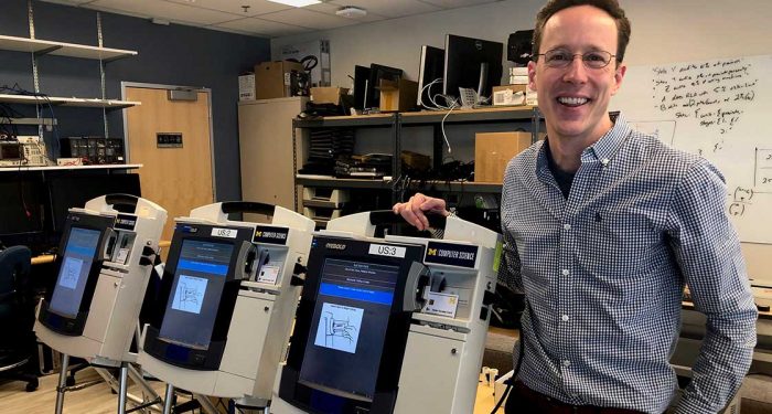 Alex Halderman stands to the right of three voting machines. He leans against the machine closest to him and smiles.