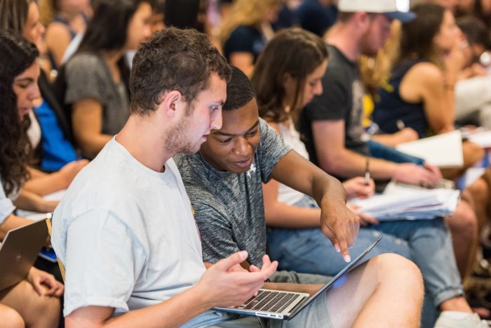 Two men discuss something over a laptop in a lecture hall. 