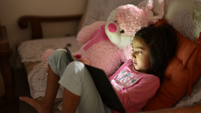 Girl sits on sofa using tablet, a pink teddy bear next to her
