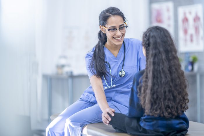 An Ethnic girl is indoors in a hospital. She is getting ready to have a checkup. She is holding a teddy bear for comfort. Her doctor is trying to relax her.