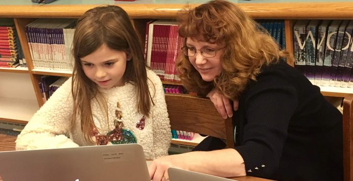 A young girl and a women look at a laptop together. They are sitting in front of a bookcase.