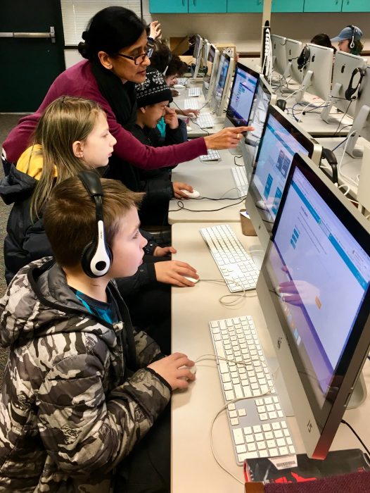 Kids work on a row of Mac computers. An adult leans over and points at a computer screen.