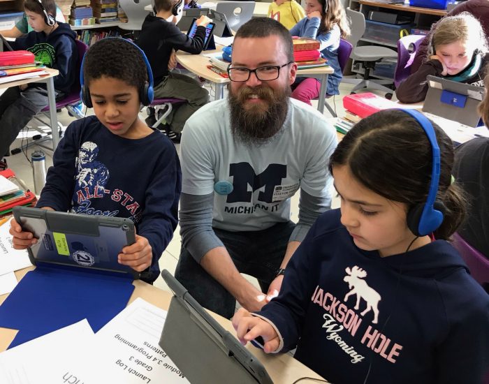 A boy and a girl work on iPads with headphones on. A man sits between them, smiling at the camera.