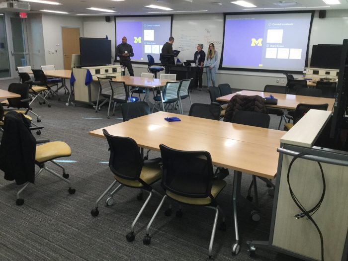 A view from the back of the team-based-learning classroom at 1280 LSA, with two large projection screens and four individuals at the front of the room.