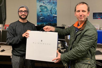 Bearded man wearing glasses and smiling accepts computer box from a smiling older man.