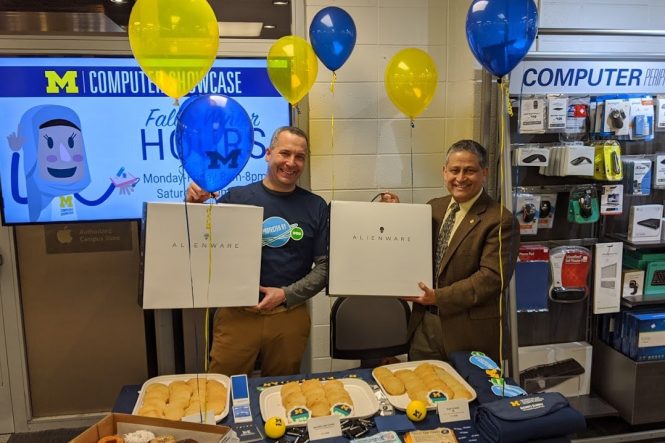 Sol Bermann and Ravi Pendse standing behind table holding laptop boxes. They stand behind a table covered in balloons, treats, and SWAG.