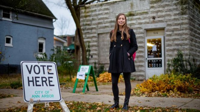 young blond woman wearing a blue coat stands in front of a building next to a "Vote Here" sign.