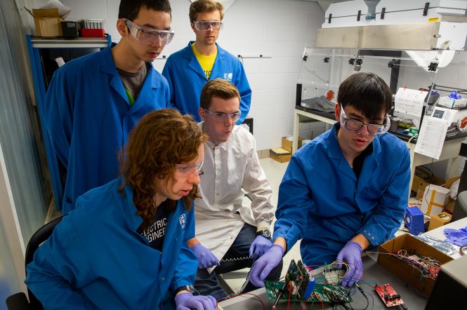 (Photo of five people in lab coats working on a piece of technology connected with many wires.)