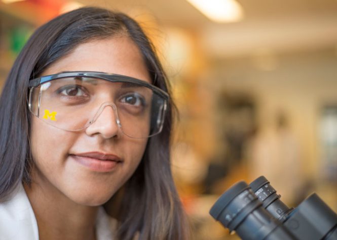 (Photo of a woman wearing lab goggles while sitting in front of a microscope.)