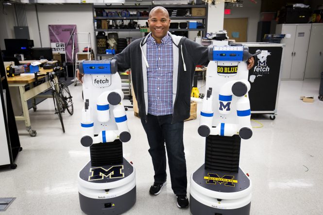 (Chad Jenkins standing with his arms around two robots that almost reach his shoulders standing upright.)