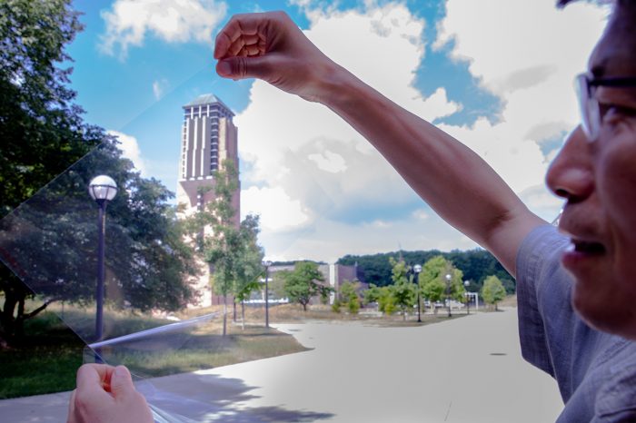 (Image of Jay Guo holding a sheet of clear conductor on the College of Engineering North Campus. Through it, a lamp and tall building are visible.)