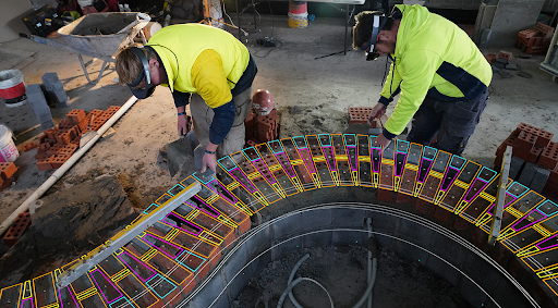 (Photo of two men wearing virtual reality headsets leaning over a virtual machinery belt in a workshop with bricks, a wheelbarrow, and concrete dust on the floor.)