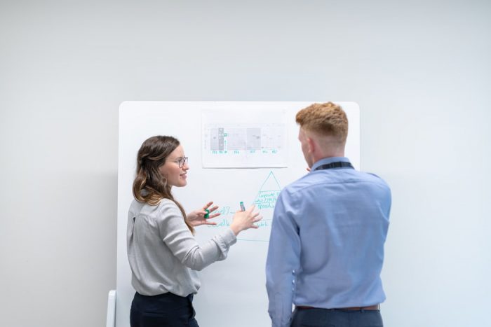 (Photo of a woman explaining something on a whiteboard to a man standing beside her.)