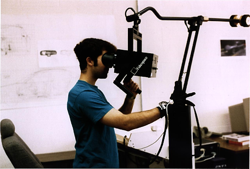 (Photo of a man looking into a rectangular black box, which is at eye level, with handles underneath, one of which he is gripping.)