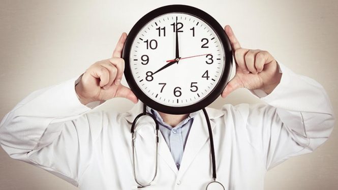 Man in lab coat with stethoscope holds clock in front of his face.