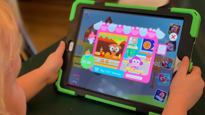 (iPad screen showing a child's game with a cartoon dog working a food stand and a monkey sitting at a desk with books. The iPad is held by a child's hands.)