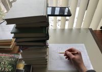 smartphone on a stack of books, pointing down toward a hand writing on a piece of paper