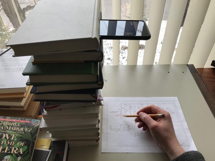 smartphone on a stack of books, pointing down toward a hand writing on a piece of paper