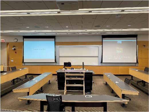 A classroom at the Ross School of Business that includes an instructor's desk in the middle, rows of desk on either side of instructor's desk, and two screens that display classroom content. 