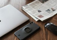 black smartphone on table next to laptop and newspaper