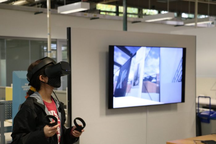 female student wearing VR headset using hand conrollers, monitor in background