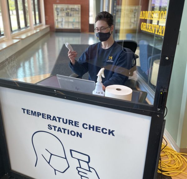 A masked student worker sits behind a screen. They are holding a thermometer and are sitting in front of a laptop. Image by Nicholas Capul.