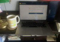 Image of a Mac laptop showing the weblogin window sitting a home desk next to U-M coffee cup.