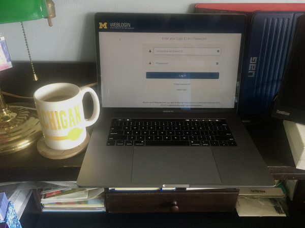 Image of a Mac laptop showing the weblogin window sitting a home desk next to U-M coffee cup.