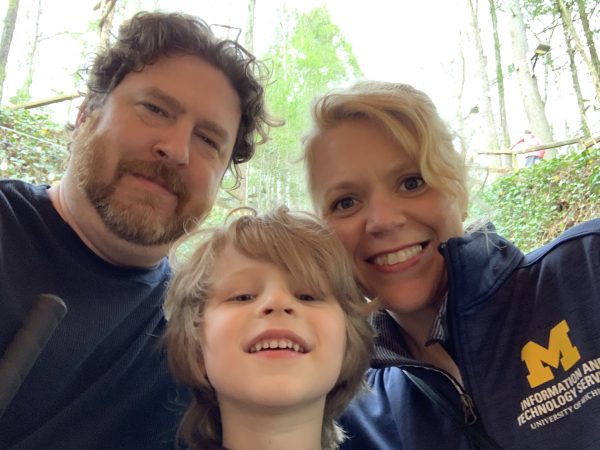 A man on the left, a woman on the right, and a boy in the middle with the woods as a backdrop.