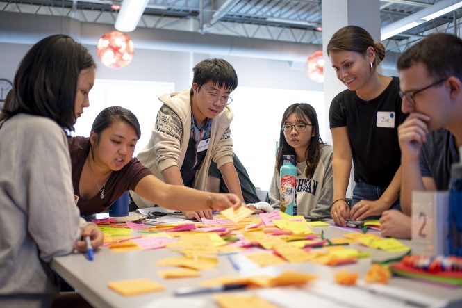A group of students gathered around a table with many sticky ntoes, analyzing a process workflow.