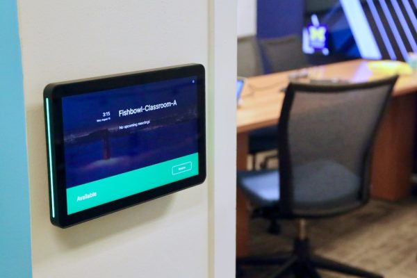 A tablet is mounted to the wall. It shows the name of the room as "Fishbowl Classroom A." There is a chair and a table in the background.