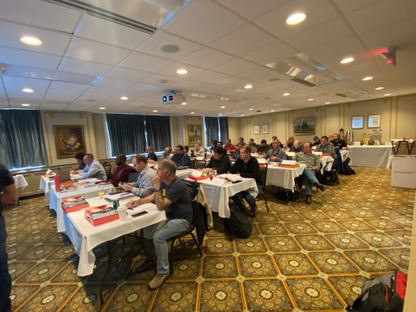 The Henderson Room at the Michigan League, set as a classroom with cloth-covered 2-person tables in a 3x5 grid. Each table has its two students facing the instructor at the front of the room, just visible to the left of frame. A snack table with dispensers and cups for coffee, orange juice, and water is at the back of the room.