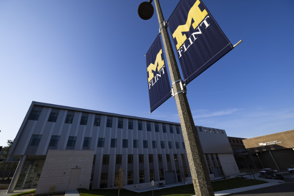 A building on UM-Flint's campus with two UM-Flint flags on a light pole out front. 