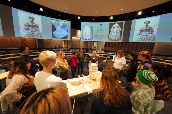 Sarah Oliver stands mid-right, wearing a crinoline and laughing with students in her classroom. Slides with images of period-piece clothing are visible on screens in the background.